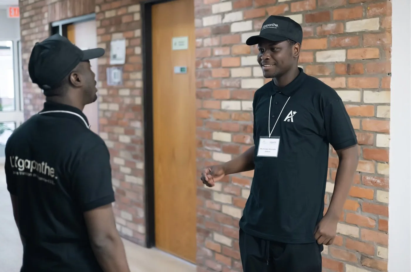 Two L'Agapanthe staff members in branded polos and caps speaking in a corridor.